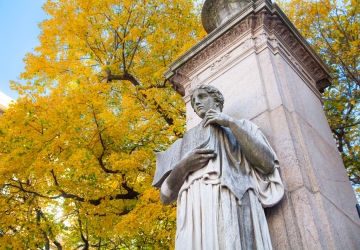 A statue of a figure holding a book in front of a tree with yellow leaves