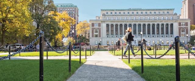A student walks on the sidewalk in front of Butler library, with a view across the Columbia campus.