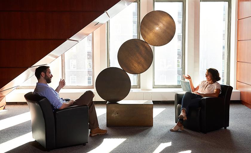 Two students sit in a lounge area by a gold sculpture with three metal discs.