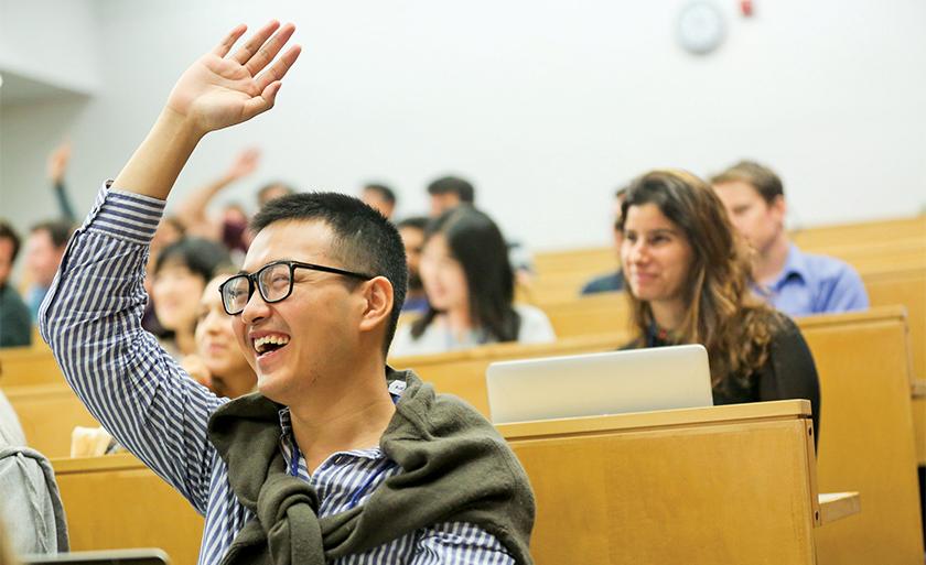 A smiling student wearing glasses raises his hand in a lecture hall.