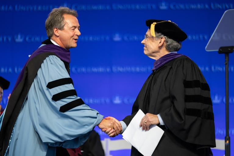 Former Montana Governor Steve Bullock and Professor Michael Gerrard dressed in academic regalia shake hands on the graduation stage