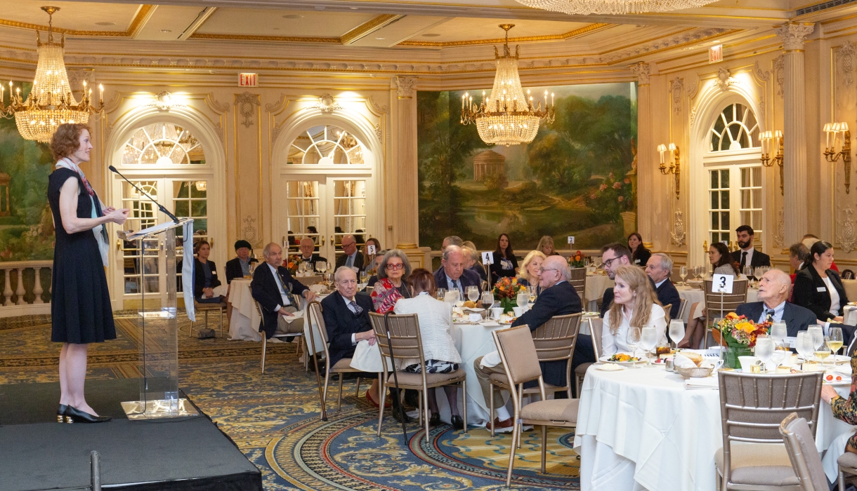 Person on a stage speaking to audience members seated at tables in a dining room