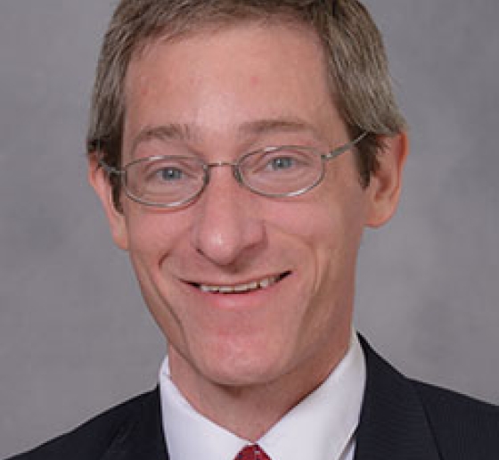 Headshot of a man wearing glasses, a white shirt, black blazer, and red tie